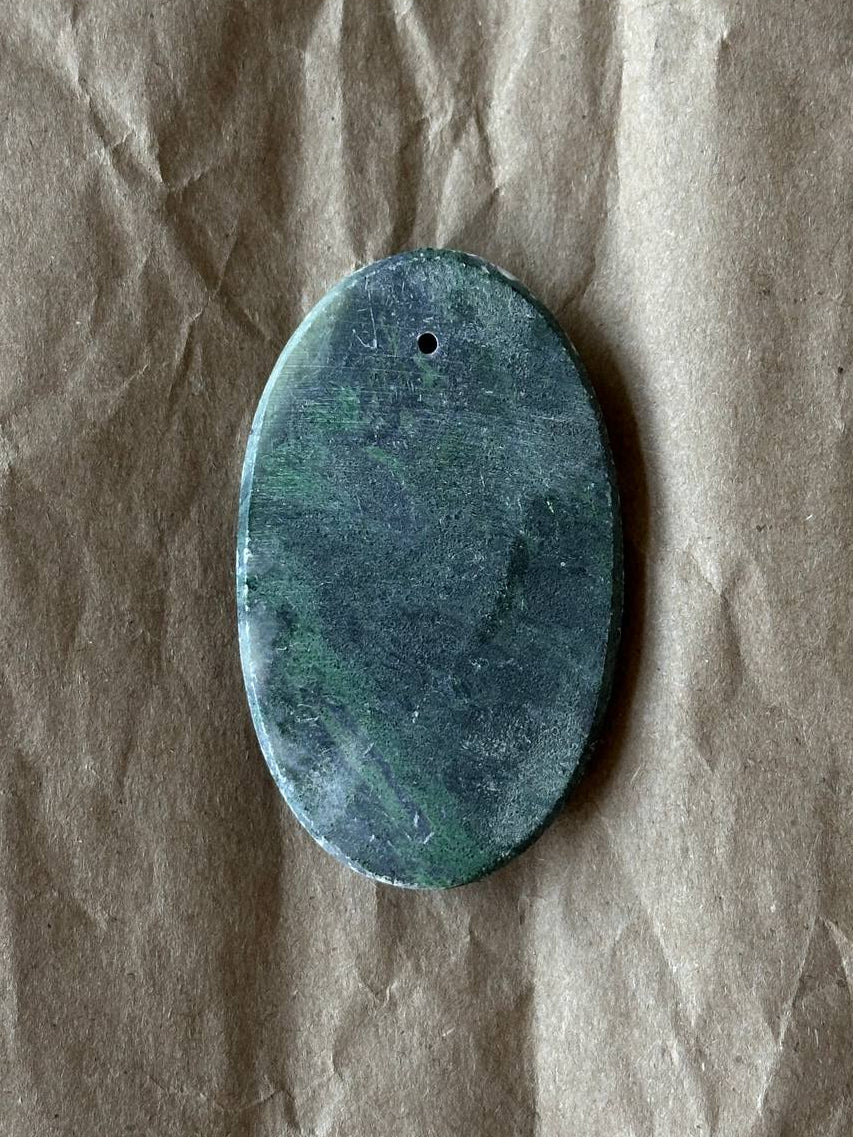 An oval-shaped green stone pendant with a small hole at the top, resting on a crumpled brown paper background.