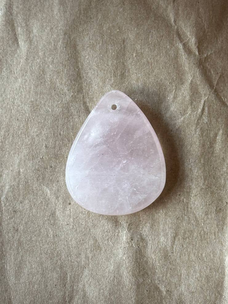 A teardrop-shaped pale pink stone pendant with a small hole at the top, resting on a textured beige paper background.
