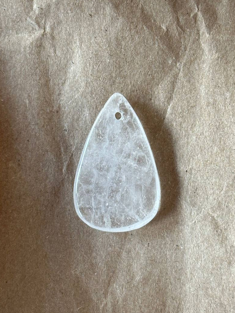 A teardrop-shaped white crystal pendant with a small hole at the top, resting on a textured brown paper background.