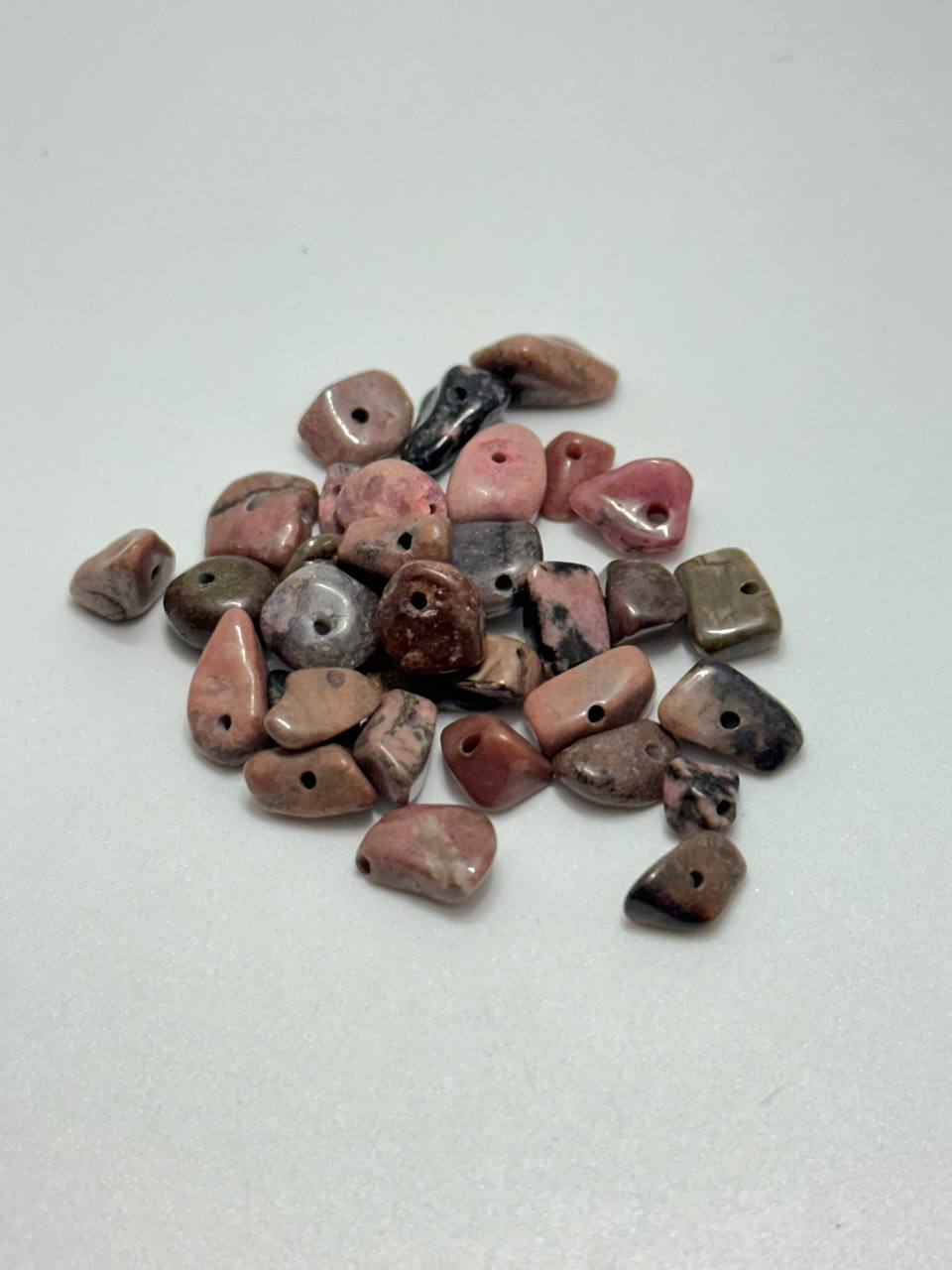 A pile of small polished tumbled stone beads with holes, showing natural marbled patterns in pink, brown, gray, and black on a light gray background.