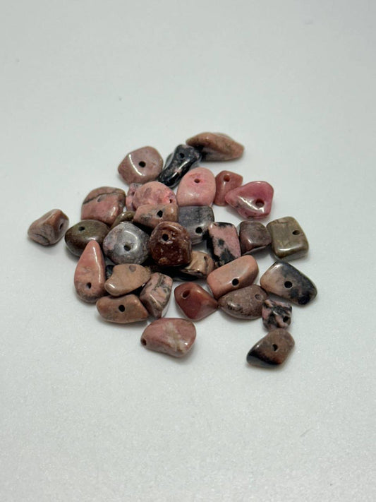 A pile of small polished tumbled stone beads with holes, showing natural marbled patterns in pink, brown, gray, and black on a light gray background.